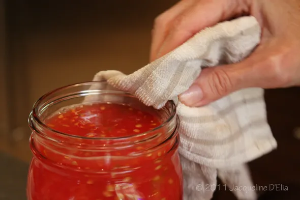 A person canning tomatoes wipes a cloth over a jar of tomato sauce.