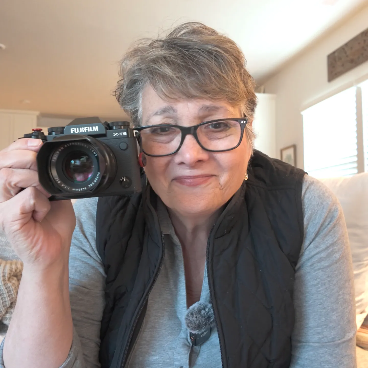 A person with short gray hair and glasses holds a Fujifilm X-T3 camera up to their face, smiling slightly, indoors.