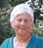 An older woman with white hair standing in front of an olive tree.
