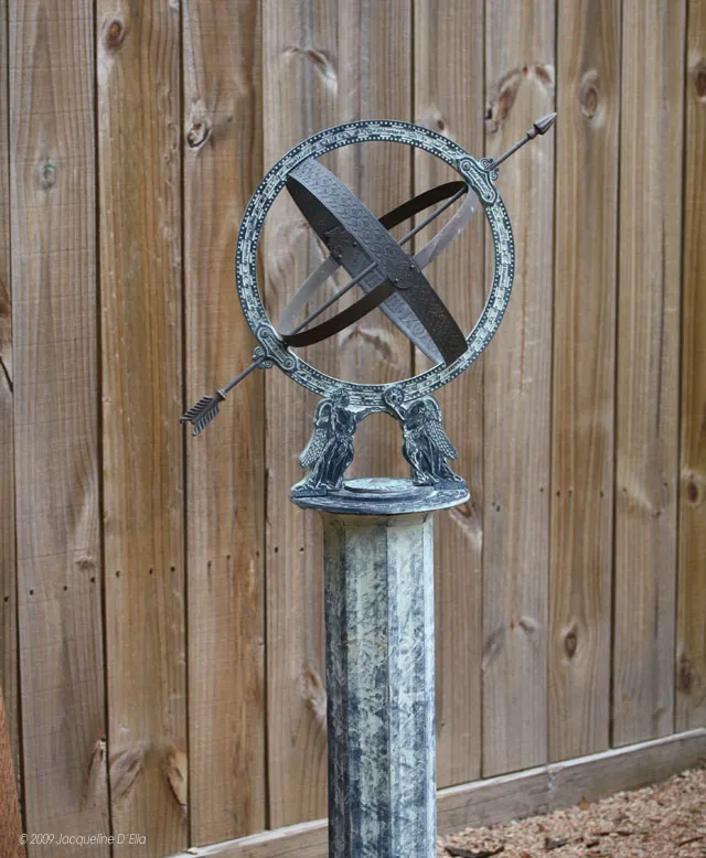 A metal sundial on a post in front of a wooden fence.
